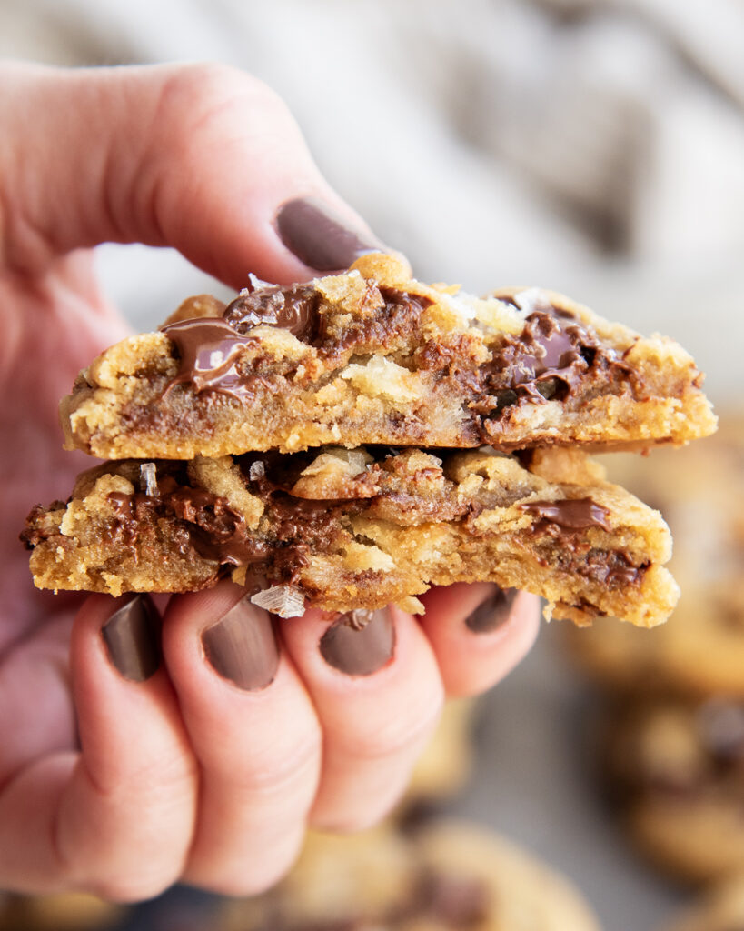 A hand holding two halves of a gooey chocolate chip cookie showing the middle of the cookie.