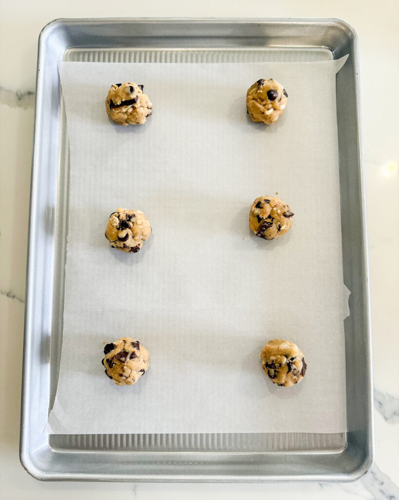 Six balls of cookie dough full of chocolate chips and toffee pieces on a baking sheet.