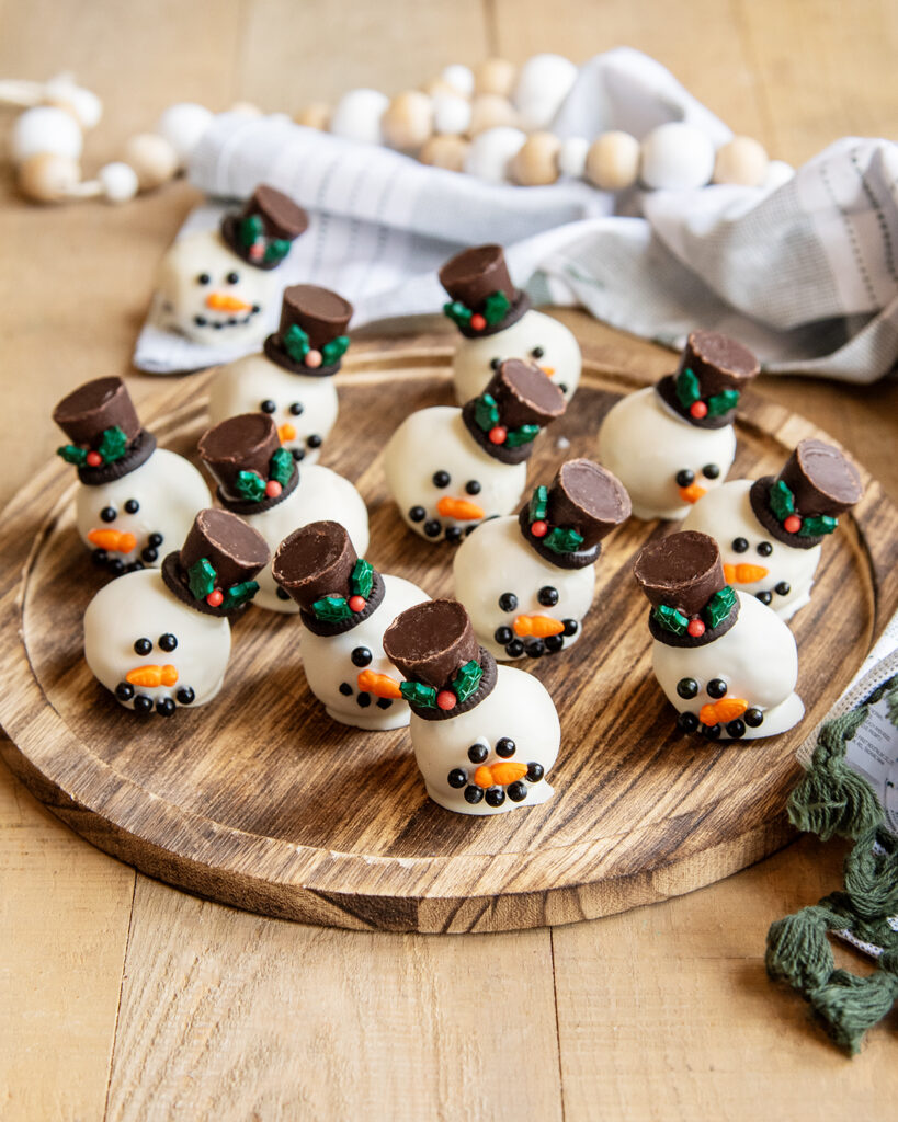 Cookie Dough balls dipped in white chocolate and decorated to look like a snowman with candy eyes, a sprinkle nose and a rolo hat, on a wooden board.