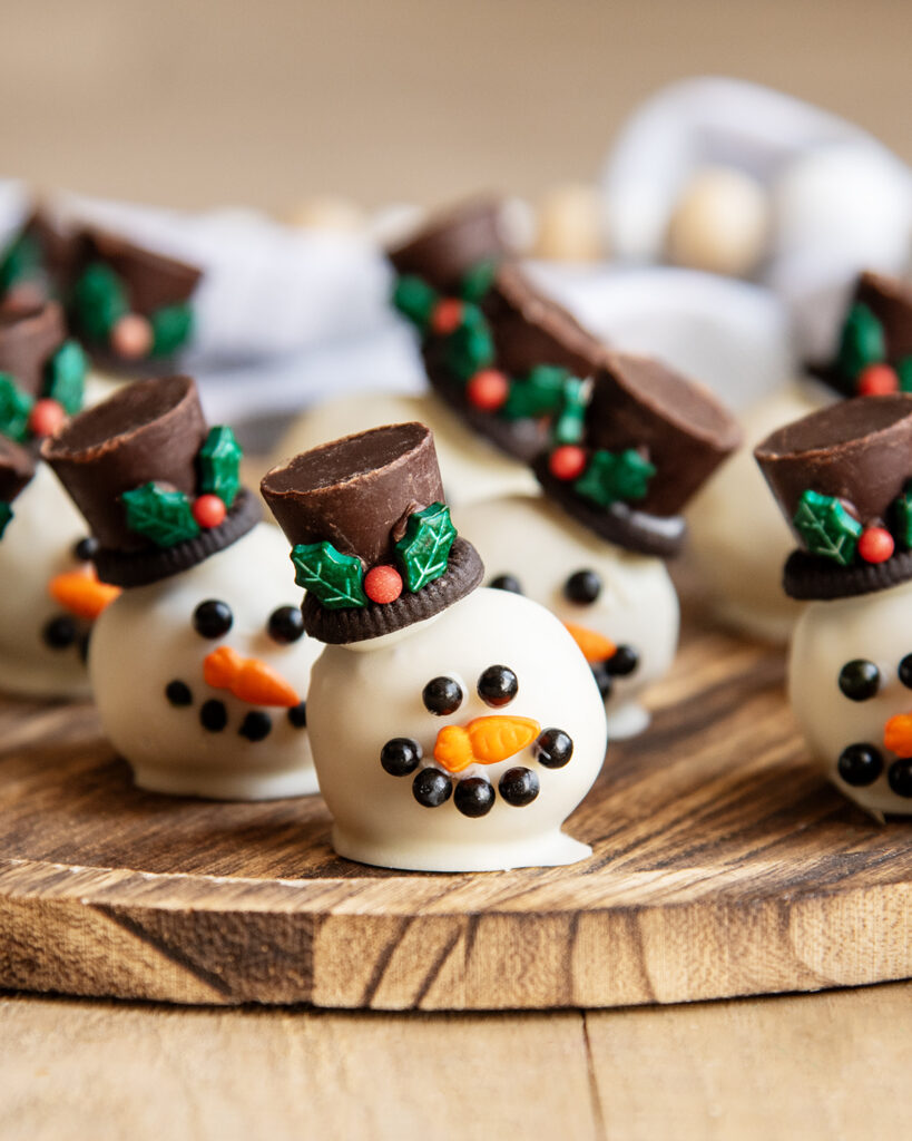 Cookie Dough balls dipped in white chocolate and decorated to look like a snowman with candy eyes, a sprinkle nose and a rolo hat. 