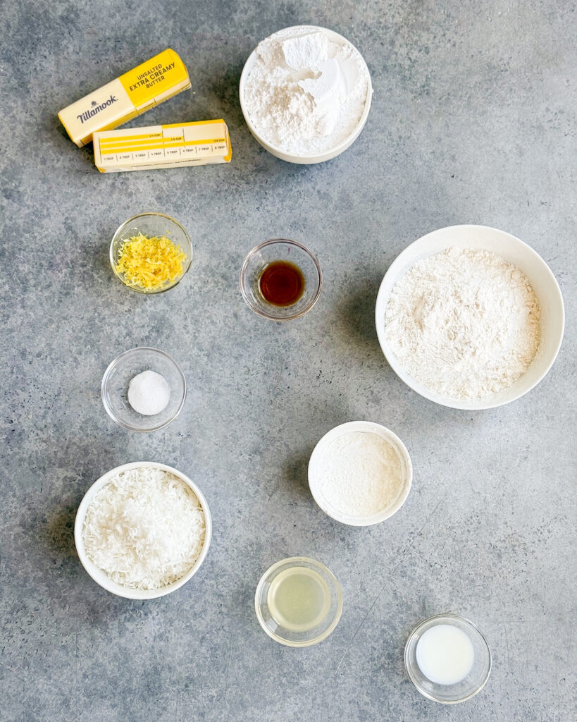 Bowls of the ingredients needed to make coconut lemon shortbread cookies, like butter, powdered sugar, flour, lemon zest, coconut, lemon juice and more. 