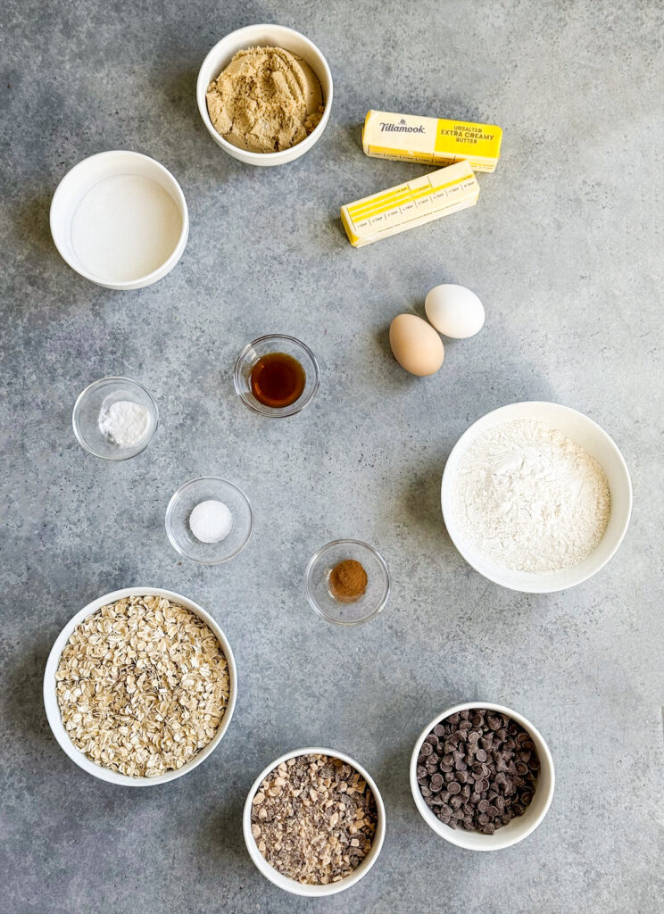 Bowls of the ingredients needed to make oatmeal toffee cookies, like oatmeal, brown sugar, butter, toffee, flour, vanilla, etc. 