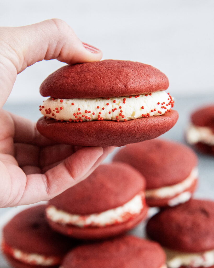 A hand holding a red velvet whoopie pie with cream cheese frosting in the middle.