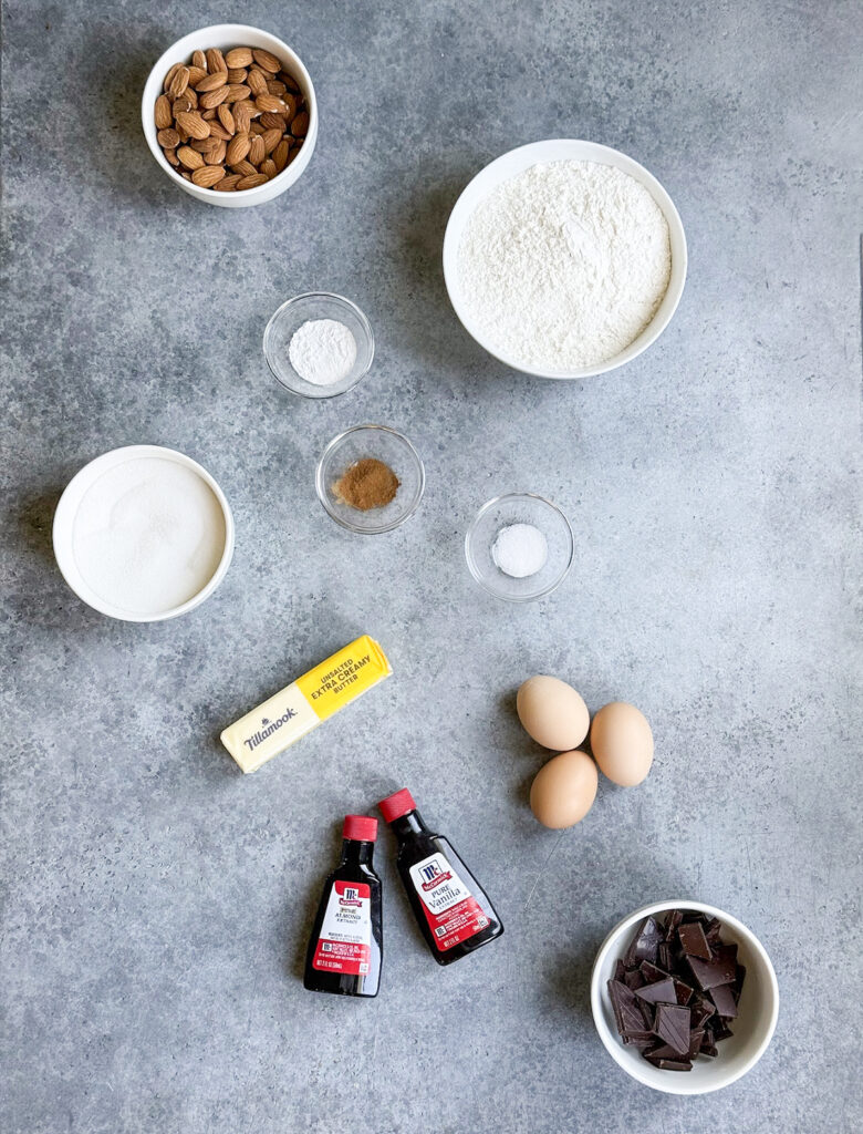 An above view of the ingredients needed to make almond biscotti, including toasted almonds, flour, sugar, butter, eggs, almond extract, and vanilla extract.