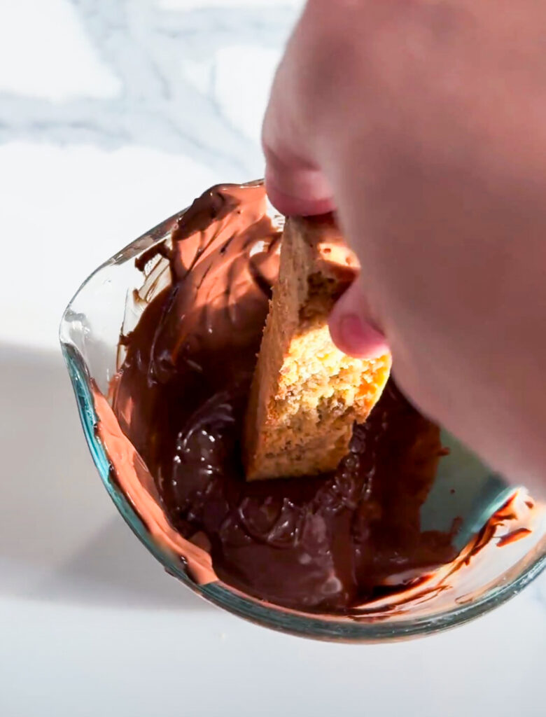 A piece of almond biscotti dipping into a cup of melted chocolate.