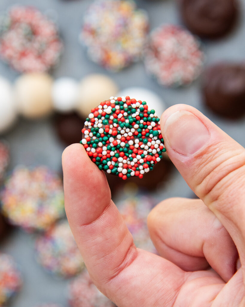 A hand holding a chocolate nonpareil showing all the round sprinkles on the bottom.