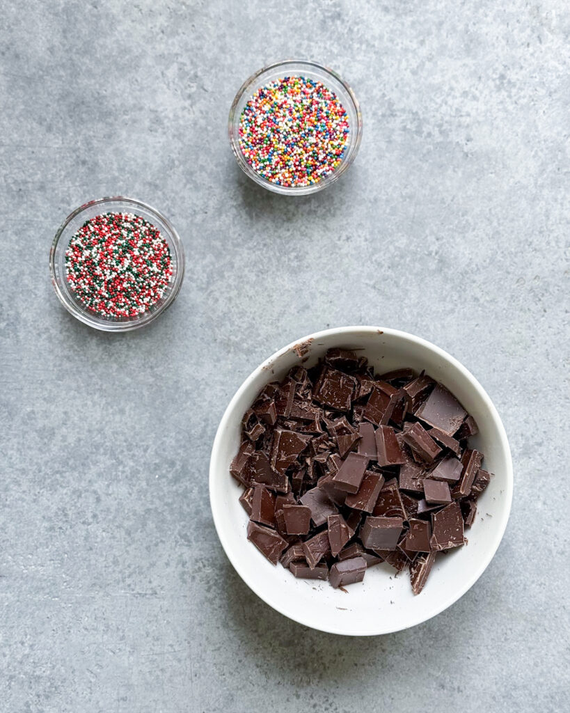 A bowl of chopped chocolate and two bowls of non pareils, one is christmas colored and one is rainbow colored.