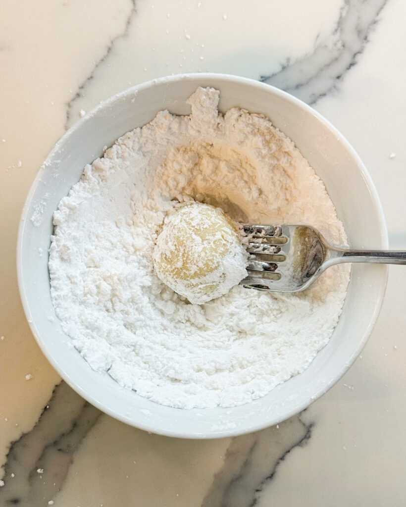A lemon cookie being rolled in a bowl of powdered sugar.