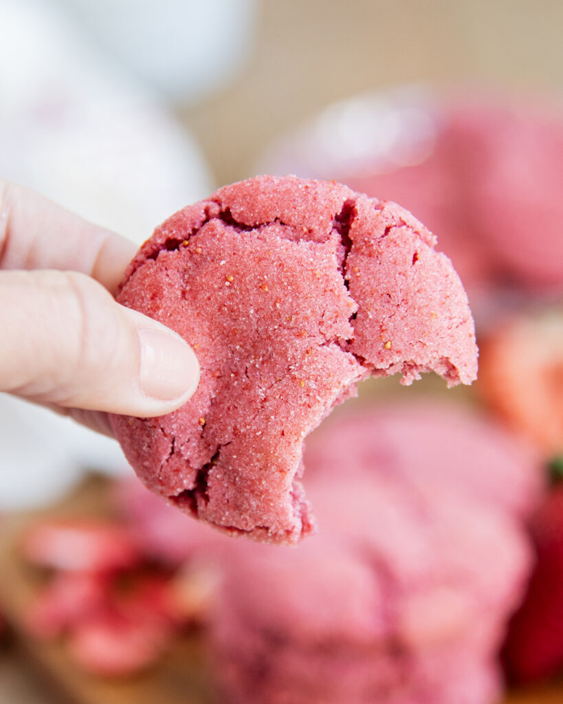 A hand holding a pink strawberry sugar cookie with a bite out of it.