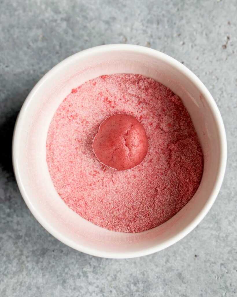 A strawberry cookie dough ball dipped in a bowl of strawberry sugar.