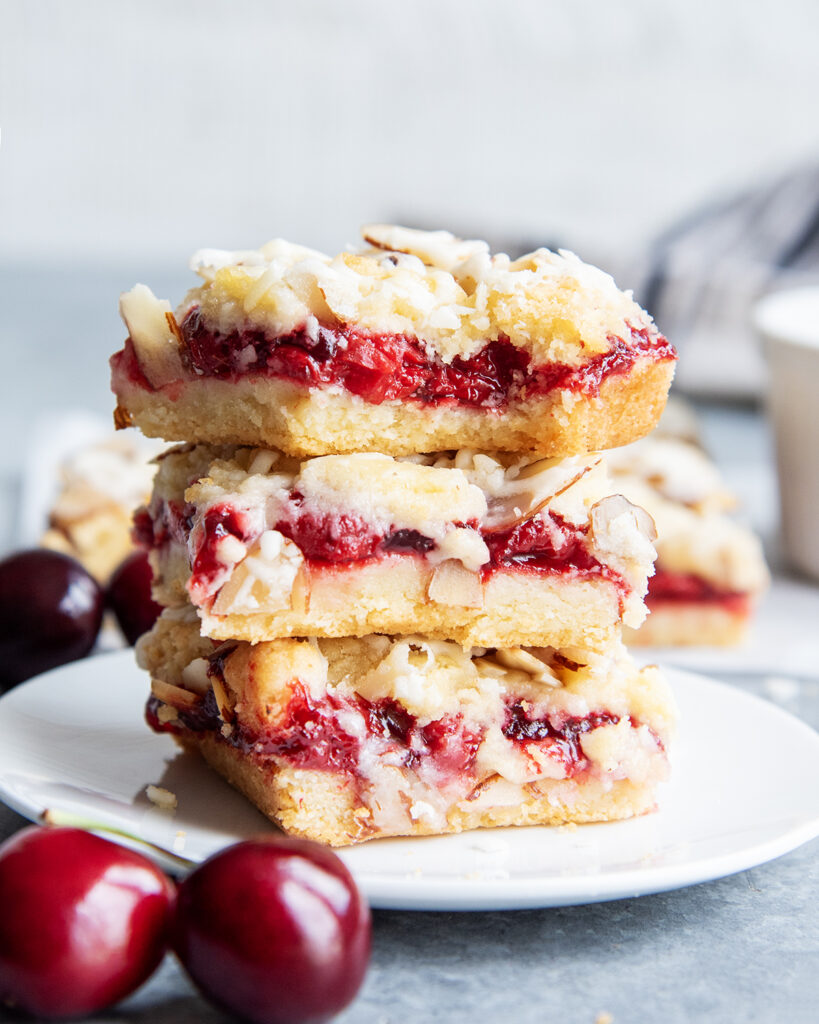 A stack of three cherry cookie bars with slivered almonds and an almond glaze on top. The top bar has a bite out of it.