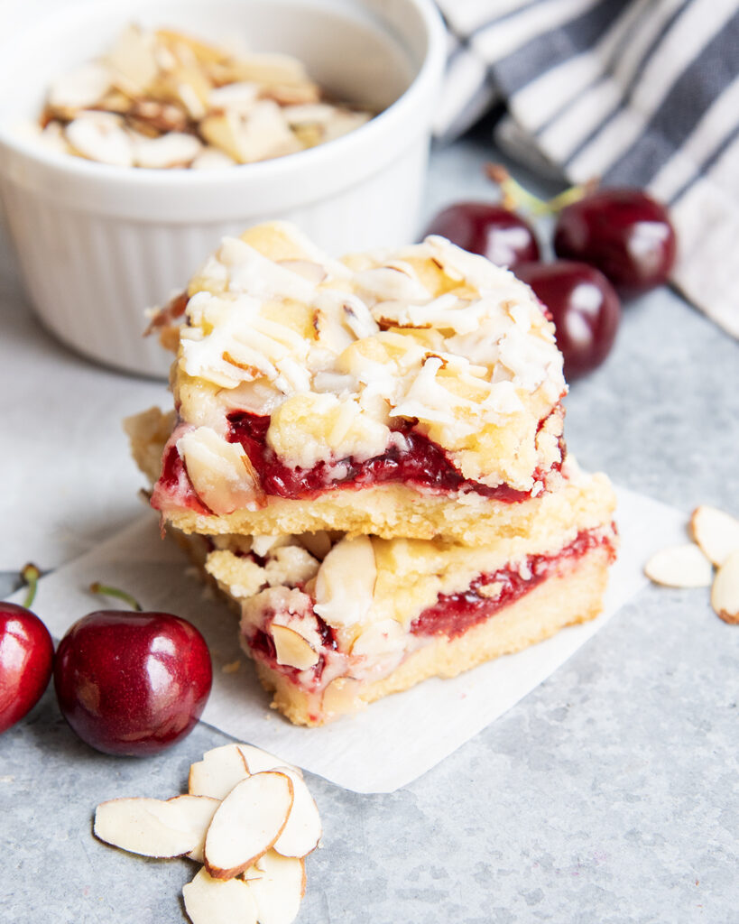 A stack of two cherry almond bars next to fresh almonds, and a bowl of slivered almonds.