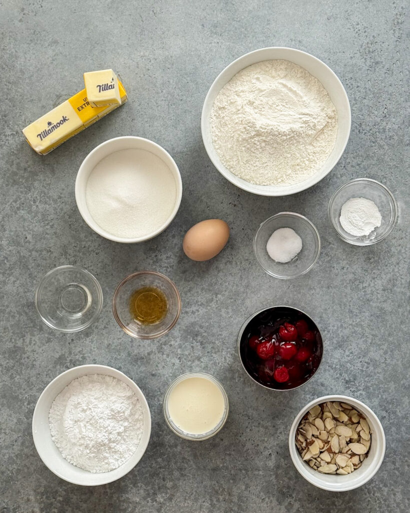 Ingredients needed to make cherry almond bars in small bowls including cherry pie filling, flour, butter, sugar, salt, slivered almonds, almond extract, and an egg.