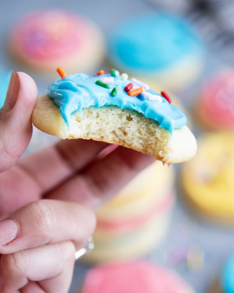 A hand holding a sour cream sugar cookie with a bite out of it showing the fluffy inside.