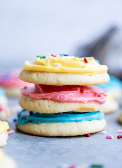 A stack of three sour cream sugar cookies, one is frosted with blue, one pink, and one yellow frosting.