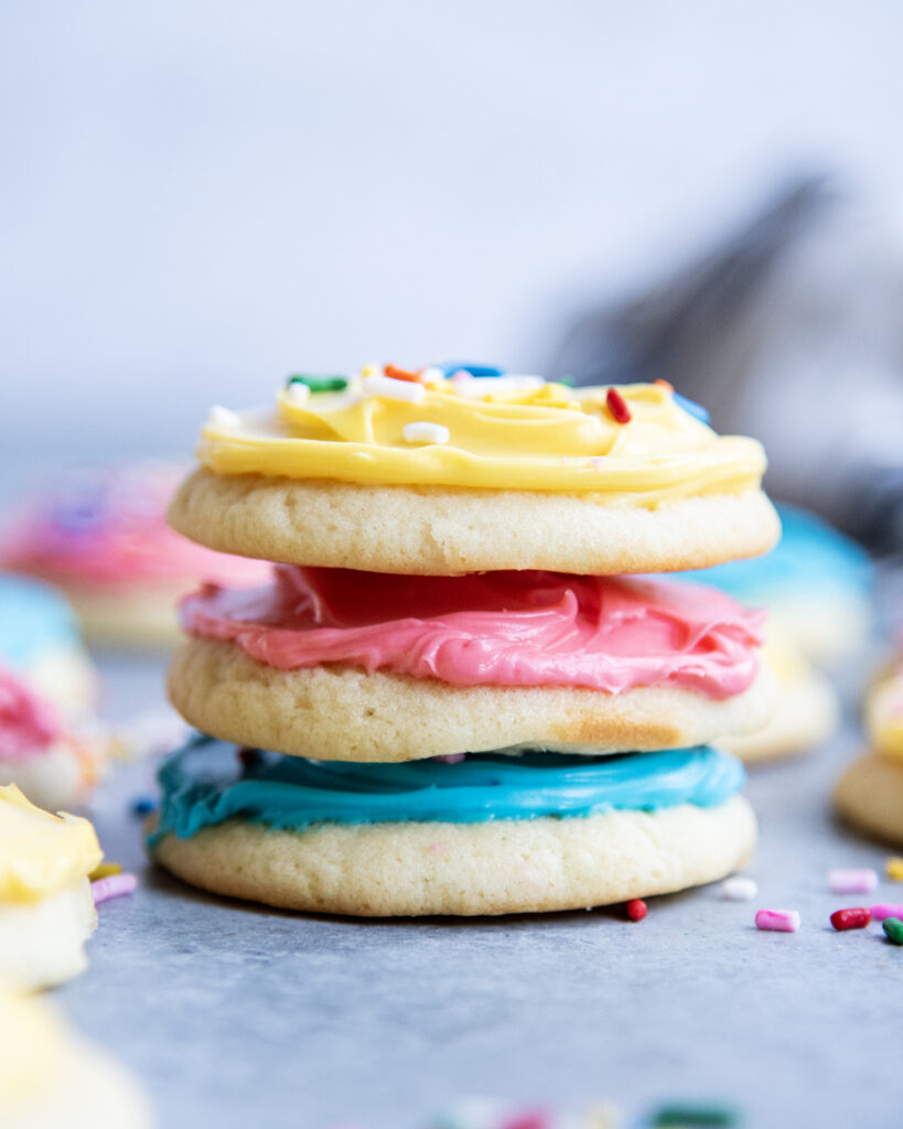A stack of three sour cream sugar cookies, one is frosted with blue, one pink, and one yellow frosting.