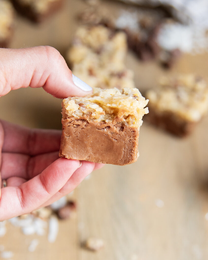 A hand holding a piece of German chocolate fudge with chocolate fudge, topped with coconut pecan frosting.