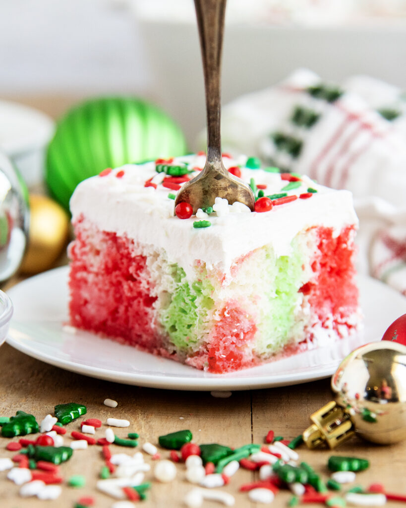 A plate of a slice of Christmas cake topped with Cool Whip, and full of red and green jello coloring, with a fork in the cake.