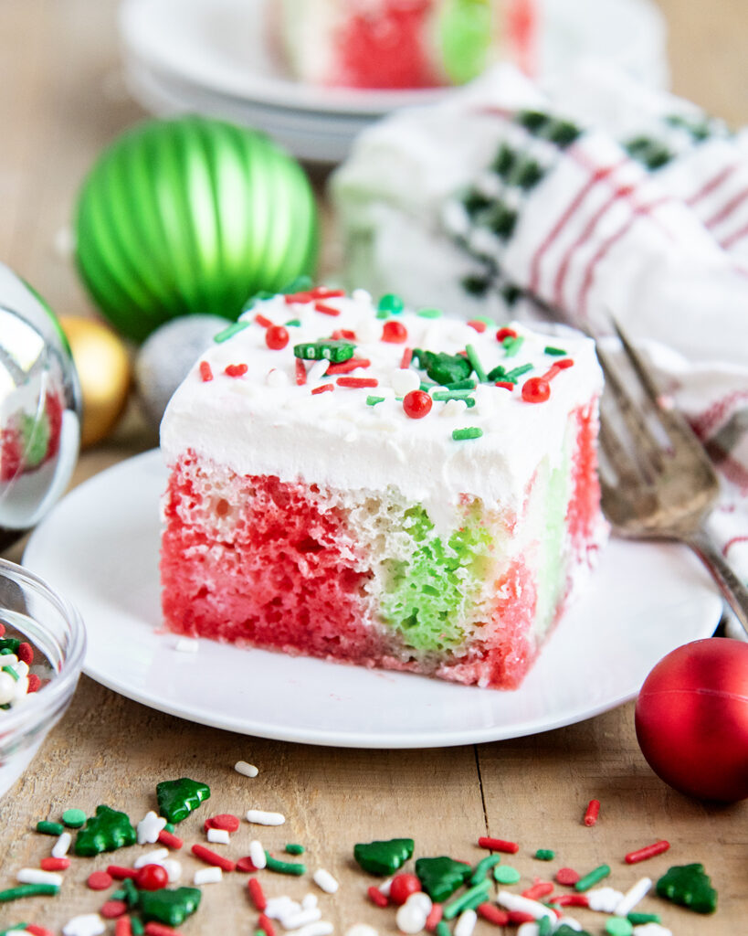 A plate of a slice of Christmas cake topped with Cool Whip, and full of red and green jello coloring.