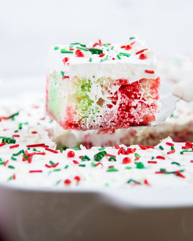 A slice of Christmas poke cake being lifted out of the pan, with red and green jello pokes.