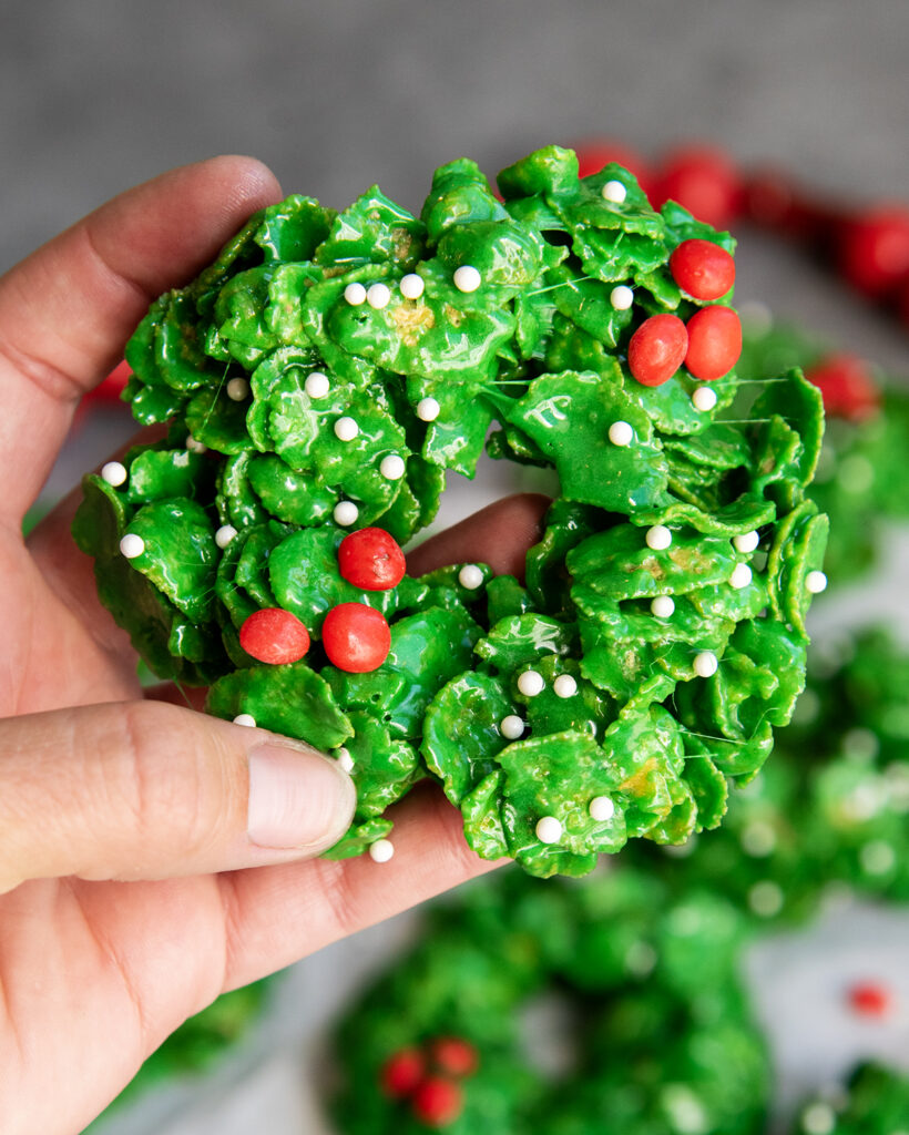 A hand holding a cornflake wreath topped with cinnamon red hots and white sprinkles.