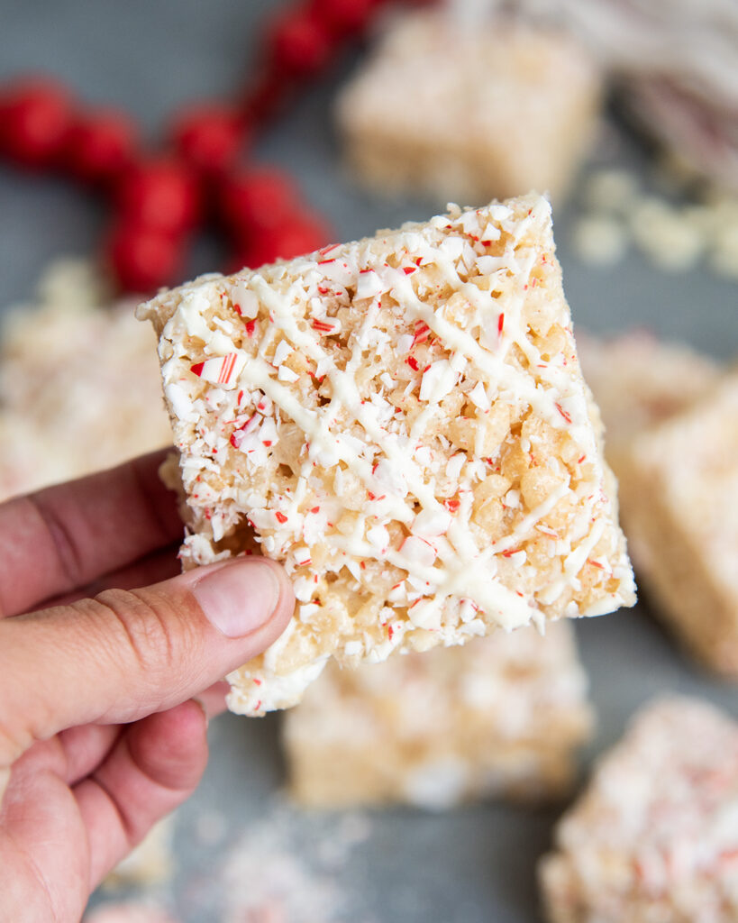 A hand holding a peppermint rice krispie treats drizzled with white chocolate and topped with candy cane pieces.