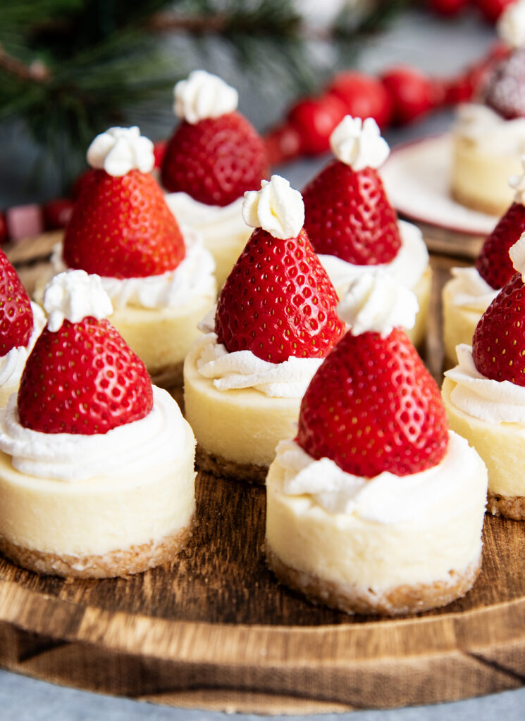 A wooden serving plate topped with mini cheesecakes with a strawberry on top.