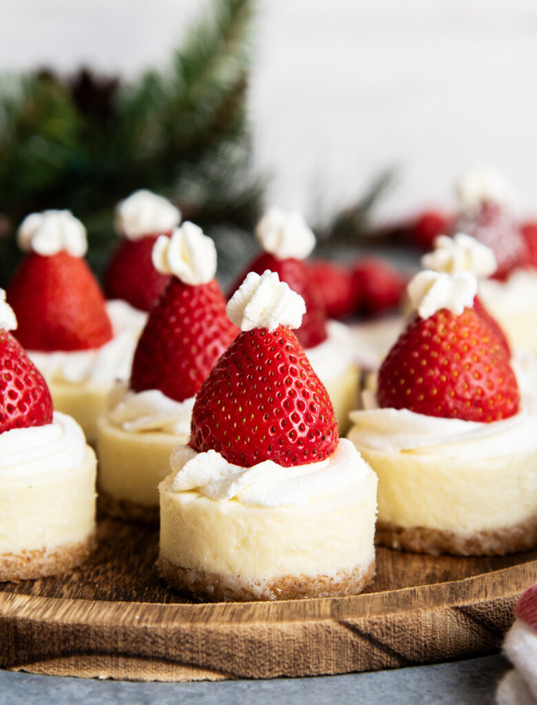 A wooden serving plate topped with mini cheesecakes with a strawberry on top.