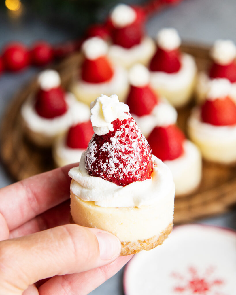 A hand holding a mini cheesecake topped with a strawberry and whipped cream.