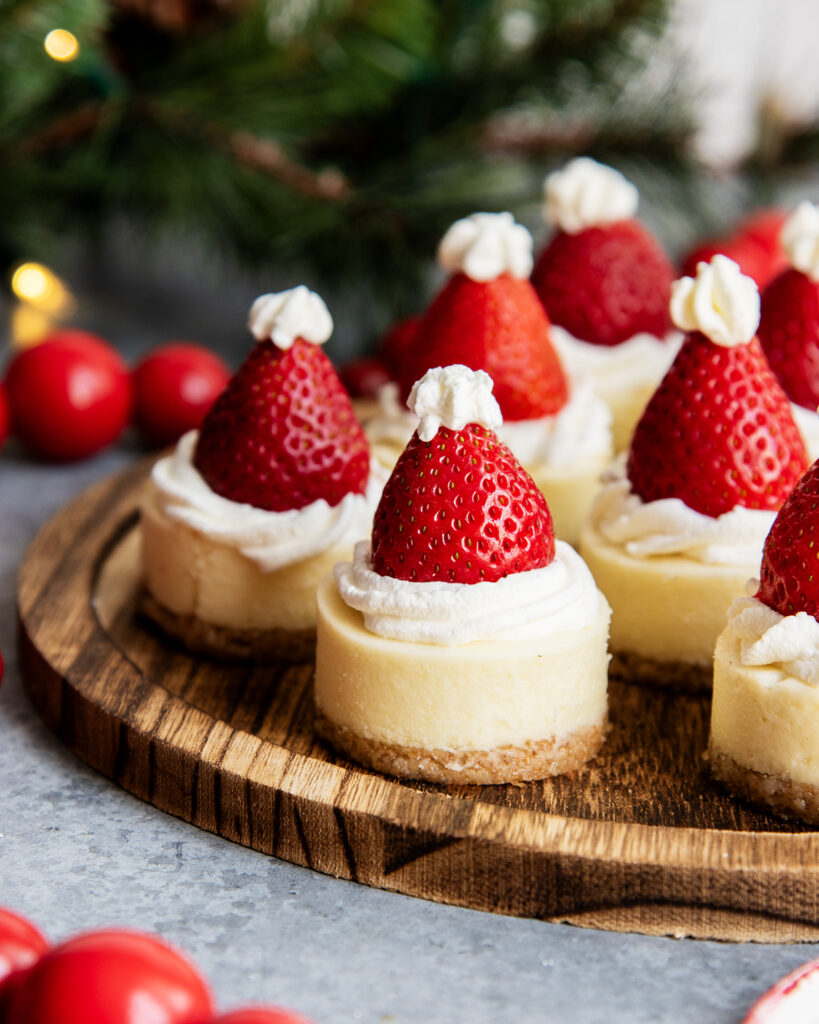 A wooden serving plate topped with mini cheesecakes with a strawberry on top.