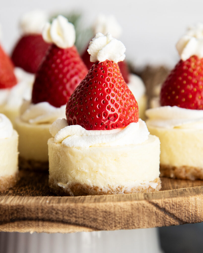 A wooden serving plate topped with mini cheesecakes with a strawberry on top.