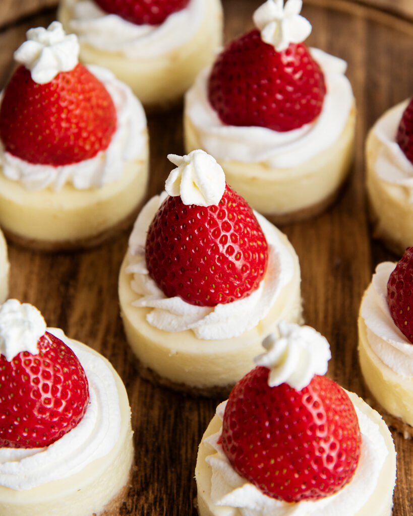 A wooden serving plate topped with mini cheesecakes with a strawberry on top.