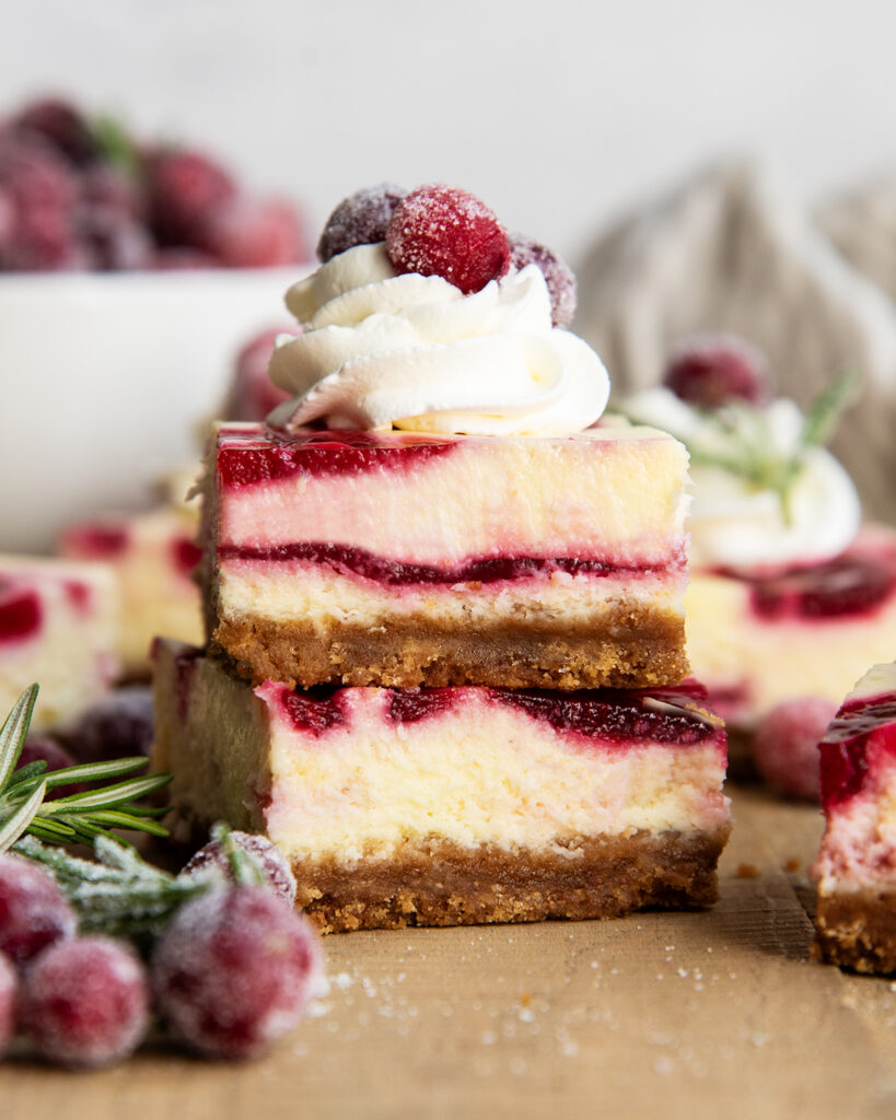 A stack of two pieces of cranberry cheesecake with cranberry sauce swirled throughout, and the top is topped with whipped cream and a candied cranberry.