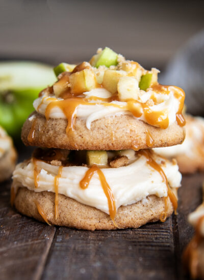 A stack of two thick cookies topped with cream cheese frosting, apple pieces, and caramel.