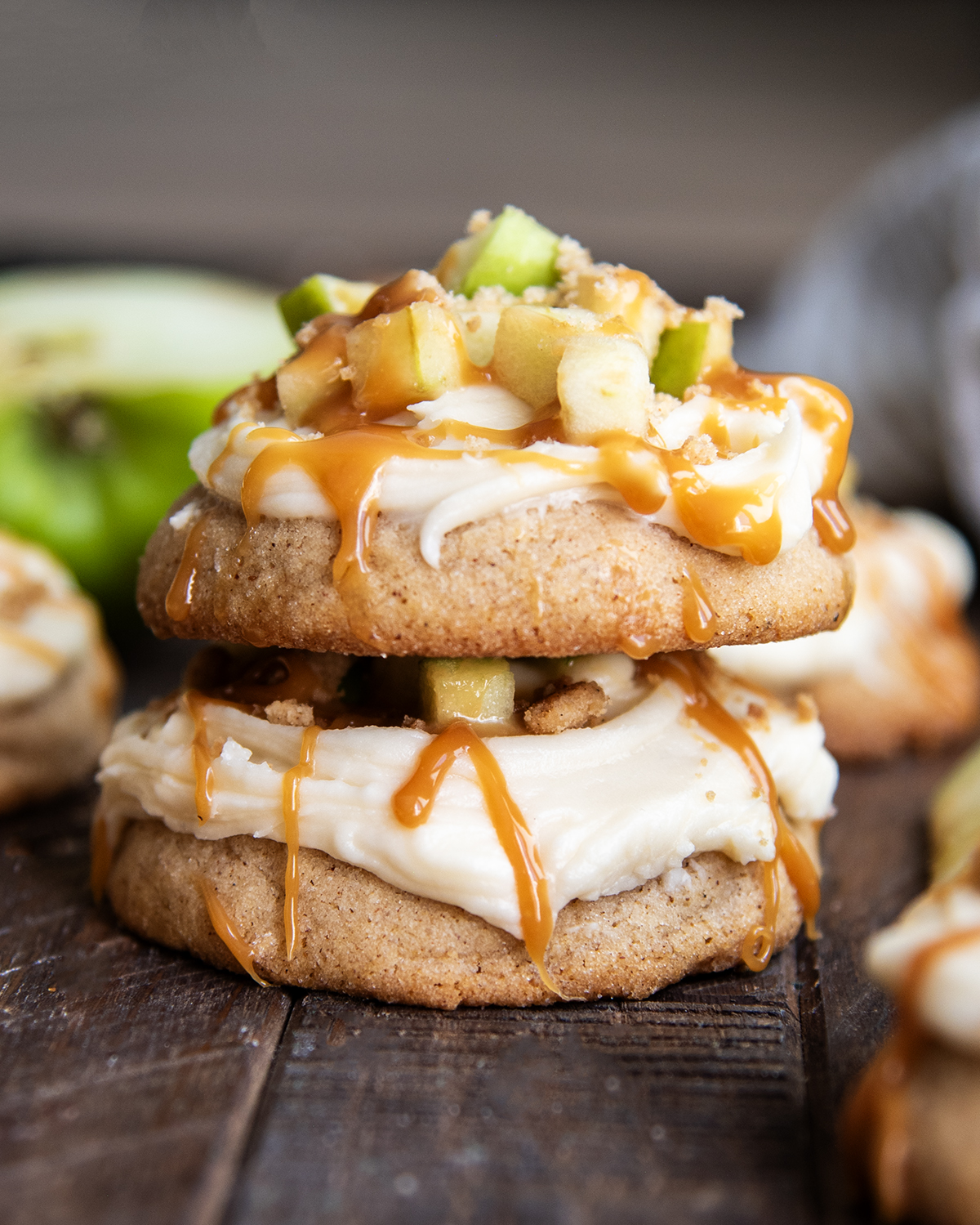 A stack of two thick cookies topped with cream cheese frosting, apple pieces, and caramel.