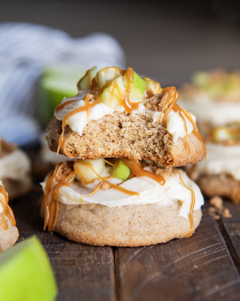 A stack of two thick cookies topped with cream cheese frosting, apple pieces, and caramel. The top cookie has a bite out of it.