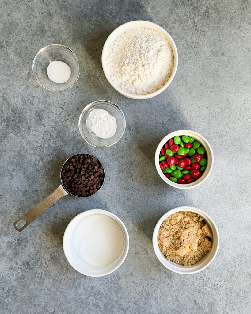 The ingredients needed to make a jar of Christmas cookie mix, all in small bowls. There is flour, m&ms, brown sugar, baking powder, and soda, etc.