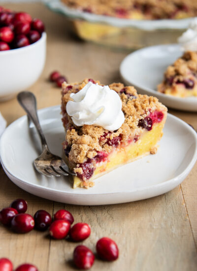 A slice of cranberry custard pie, with a crumb topping and whipped cream on top, on a white plate with a fork.