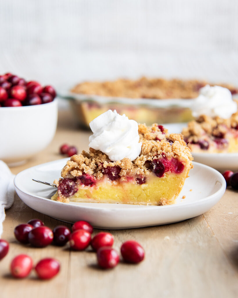 A slice of cranberry custard pie, with a crumb topping and whipped cream on top, on a white plate.
