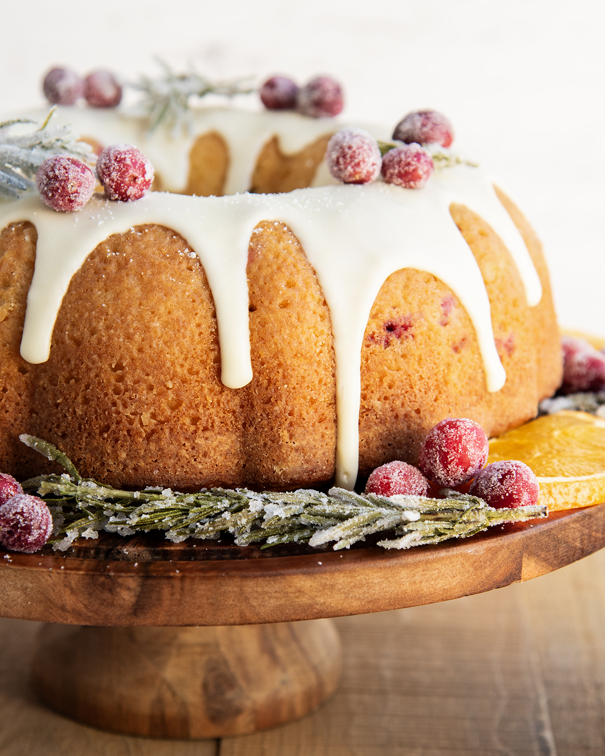 A bundt cake on a wooden stand topped with a vanilla glaze and candied cranberries.