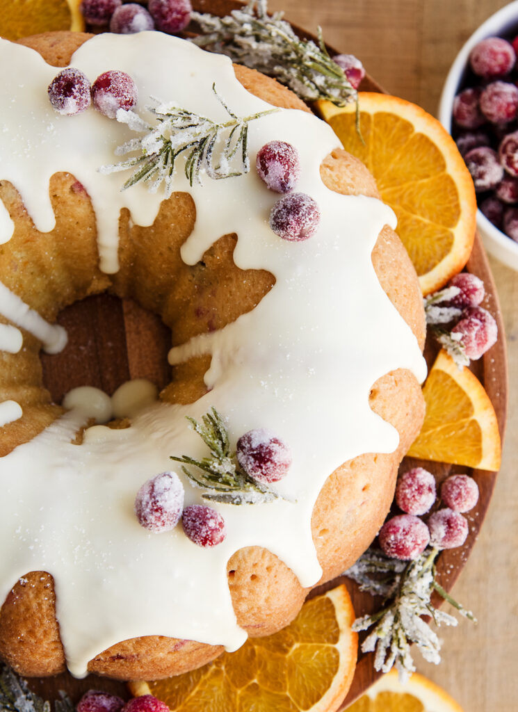 A bundt cake on a wooden stand topped with a vanilla glaze and candied cranberries.