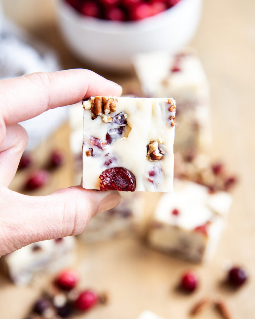 Two fingers holding a piece of white chocolate cranberry fudge.