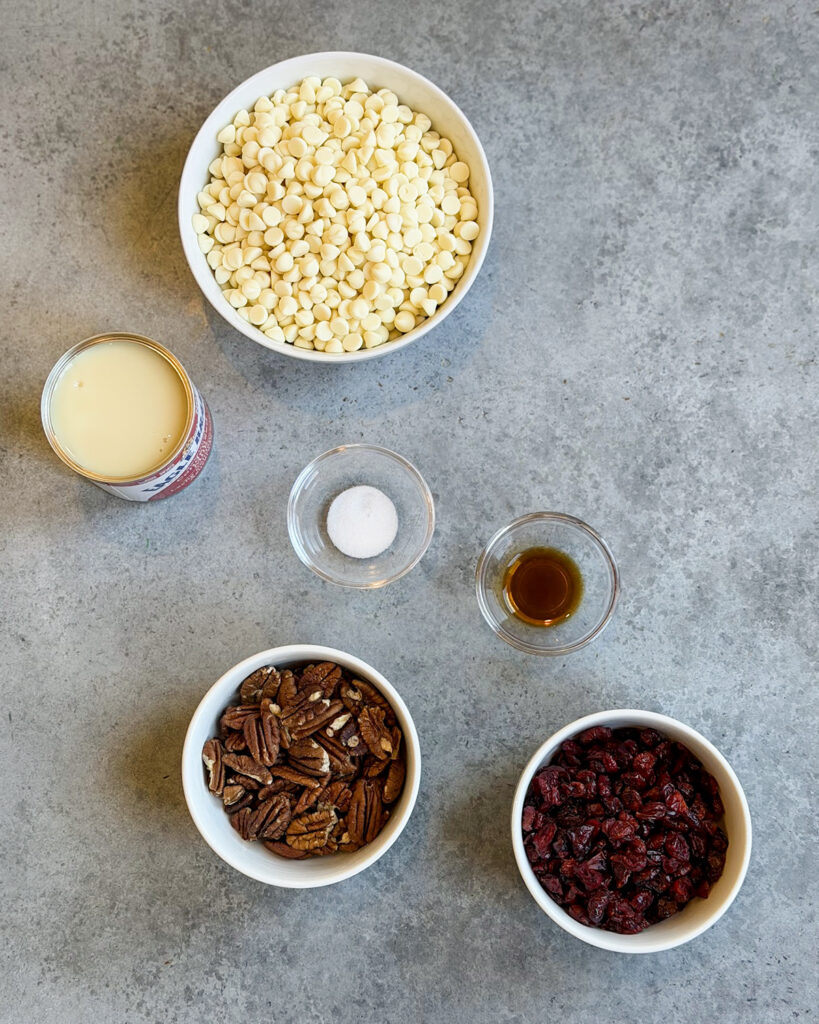Small bowls of the ingredients needed to make white chocolate cranberry fudge, like white chocolate chips, sweetened condensed milk, cranberries, and pecans.
