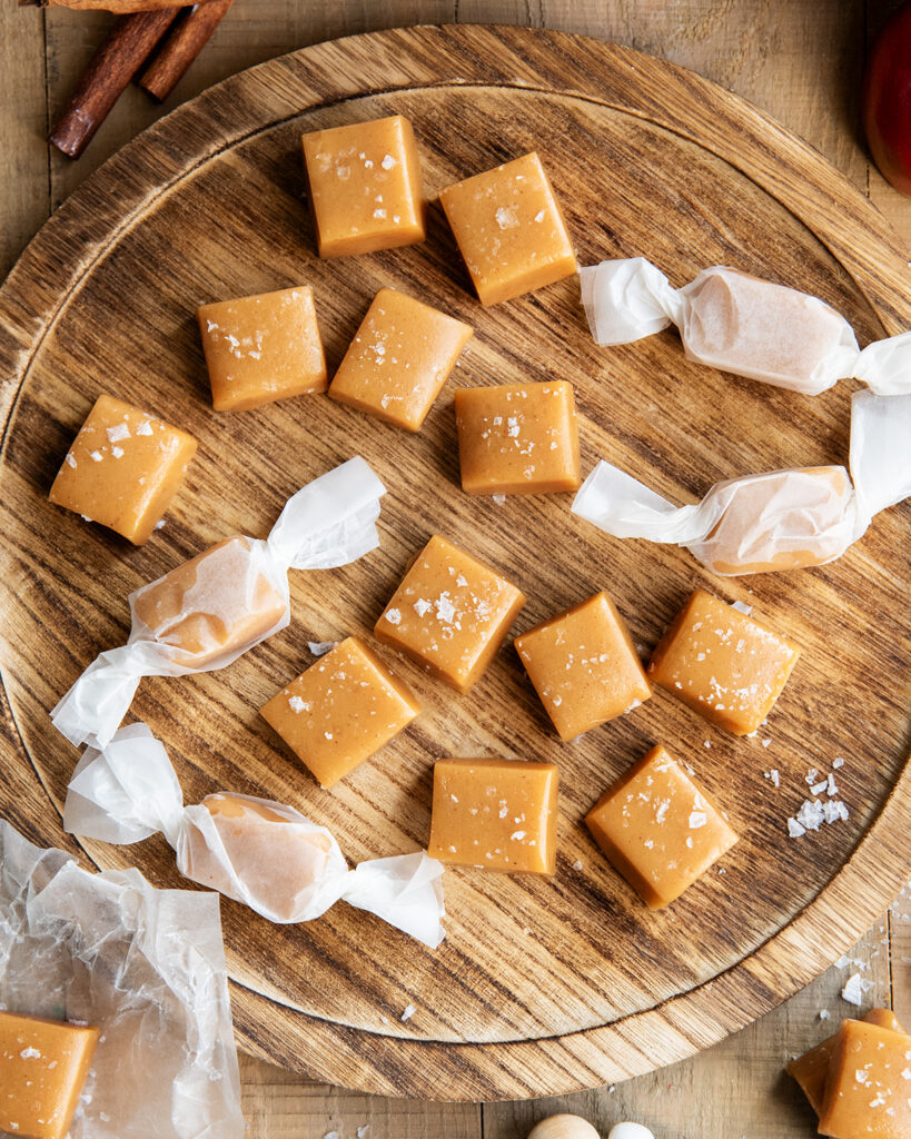 Apple cider caramels on a wooden tray, some are wrapped in parchment paper.