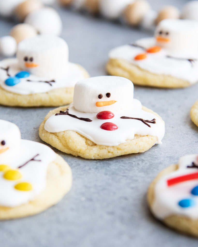An above view of melted snowman cookies decorated with royal icing, mini m&ms, and marshmallows for their heads.