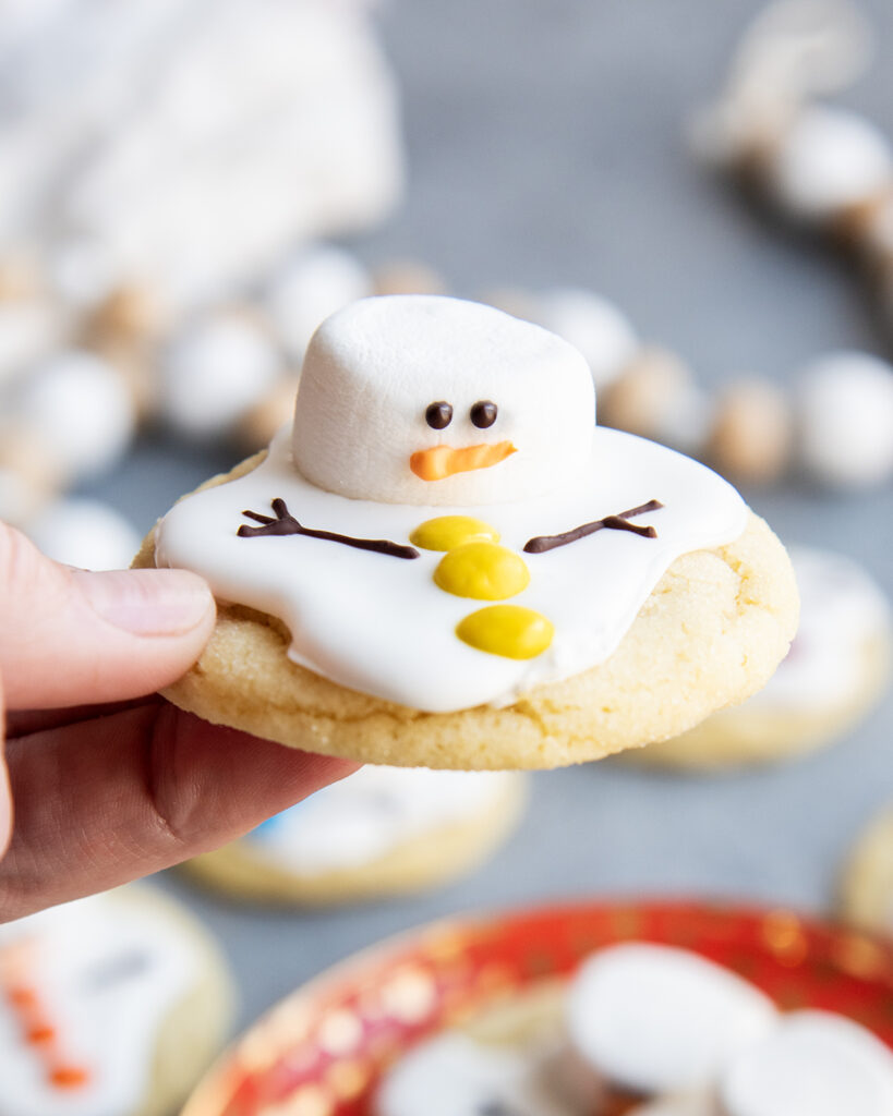 A hand holding a melted snowman cookie decorated with a large marshmallow, royal icing, and mini m&ms.