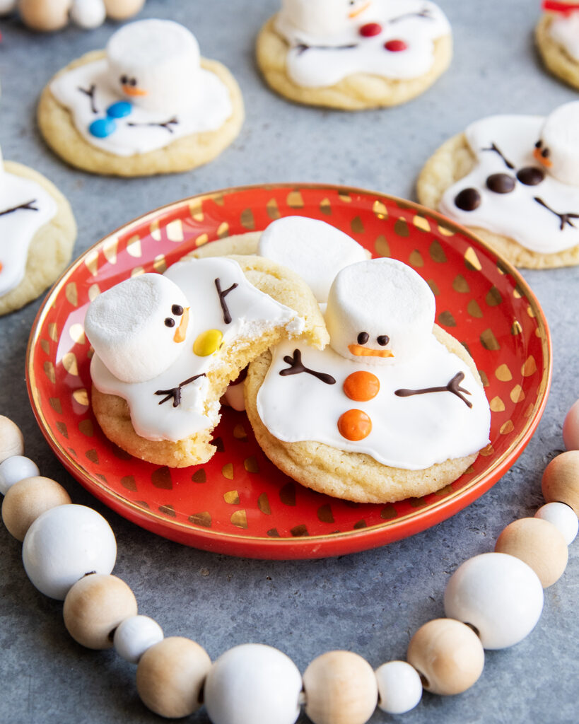 A plate of three melted snowman cookies decorated with royal icing and marshmallows. One cookie has a bite out of it.
