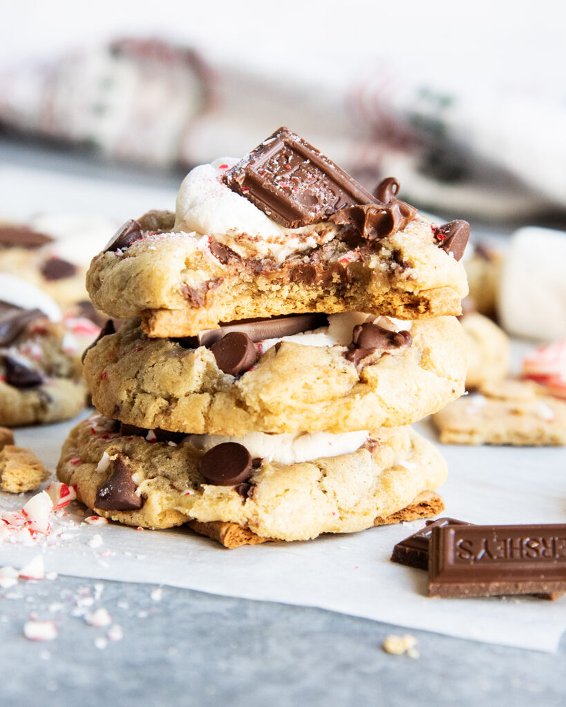 A stack of three peppermint smores cookies topped with a gooey marshmallow and chocolate piece. The top piece has a bite out of it.