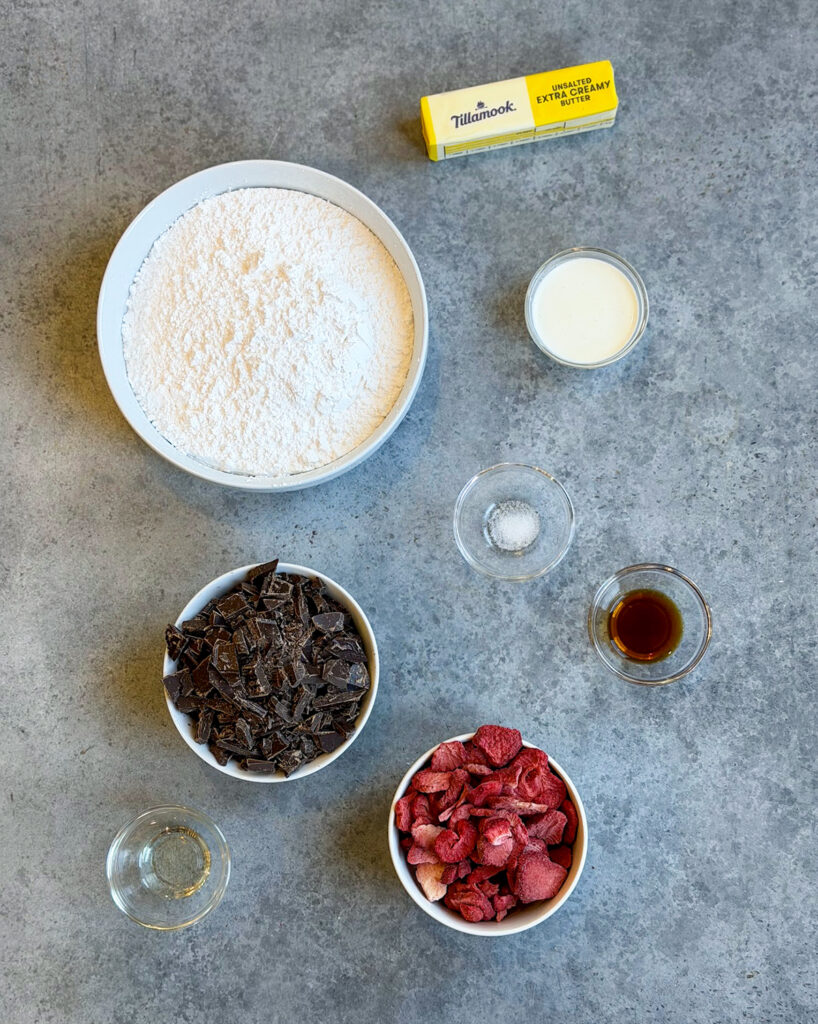 The ingredients needed to make strawberry cream chocolates in small bowls, including powdered sugar, butter, cream, chocolate, and freeze dried strawberries.
