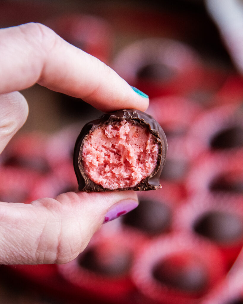 Two fingers holding a strawberry creme chocolate with a bite out of it showing the strawberry buttercream center.